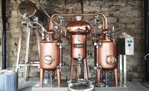 two red copper pot stills and a red copper heat exchanger in between, all on a skid distilling spirit on site