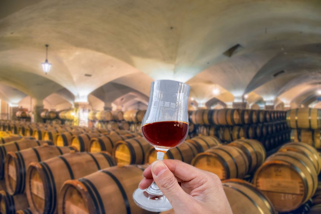 A cellar with oak barrels storing spirits and a cup of brandy hold by a hand